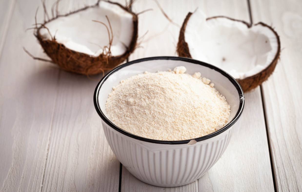Coconut flour in a bowl displayed in front of an open coconut fruit. 