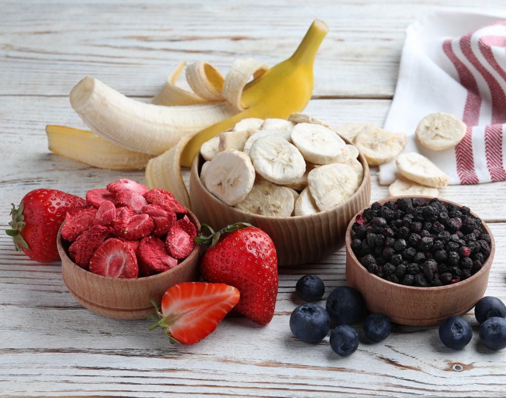 Freeze-dried banana slices, blueberries and strawberry slices each in bowls displayed with Fresh strawberry fruit, a banana and fresh blueberries.