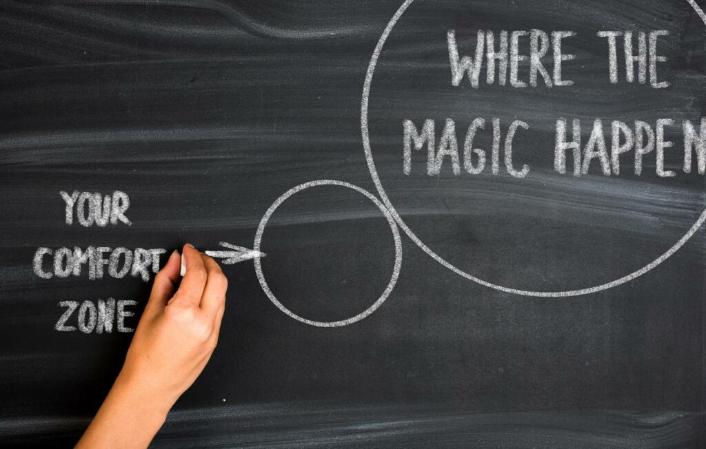 Chalk board with a hand writing with a hand writing and indicating that "where the magic happens" is outside "your confort zone"