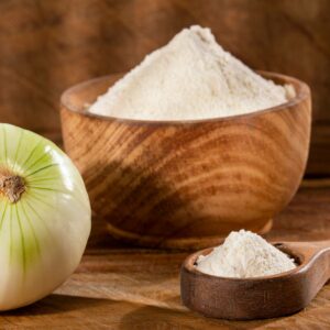 Onion powder on a wooden spoon beside an onion and a wooden bowl with more onion powder